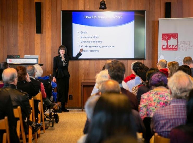 Carol Dweck gesturing toward a slide reading “How do mindsets work? Goals, meaning of effort and setbacks, challenge seeking, persistence makes better learning